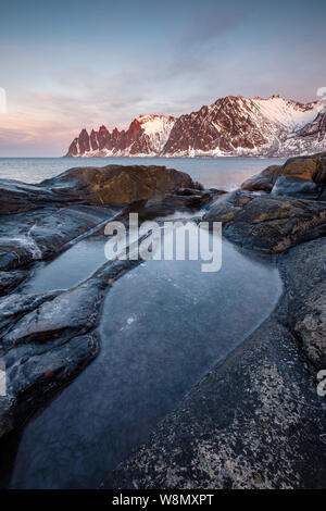 View over colorful rocks and rockpools to snowy Oksen mountains, Tungeneset Devils Jaws Senja Norway Vertical Stock Photo