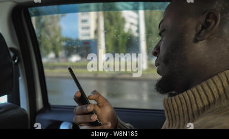 Portrait of black man is browsing smartphone. He is riding a car in rainy day in city. Afro american bearded guy is sitting in the back seat, side view. Stock Photo
