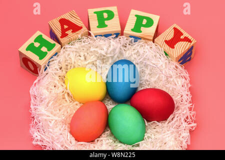 Five Easter colored eggs lie in a nest on top of cubes with the words happy on a gently pink background. Horizontal photo. Stock Photo
