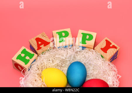 Five Easter colored eggs lie in a nest on top of cubes with the words happy on a gently pink background. Horizontal photo. Stock Photo