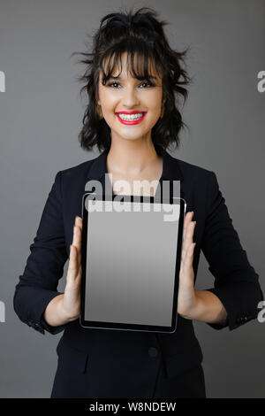 Attractive young female in stylish outfit smiling and looking at camera while standing on gray background and showing modern tablet with empty display Stock Photo