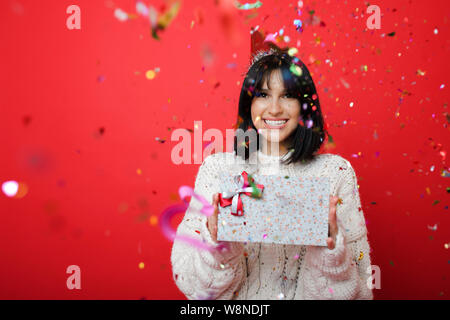 Smiling young woman holding wrapped gift box while standing in falling confetti on red background Stock Photo