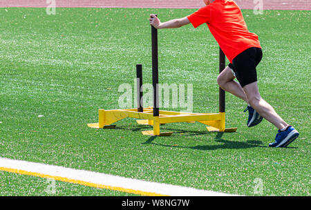 A young male runner is pushing a yellow sled on a green turf field at running camp. Stock Photo