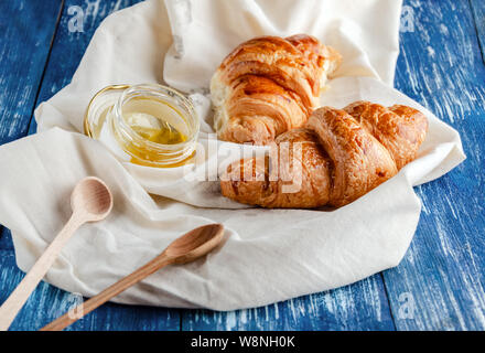 croissant with a beautiful crisp and a jar of honey on a light fabric, delicious pastries with honey and spoons in a cloth napkin on a blue background Stock Photo