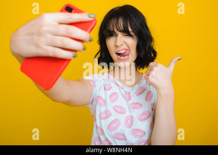 Beautiful young woman showing tongue and gesturing thumb-up while posing for selfie on yellow background Stock Photo