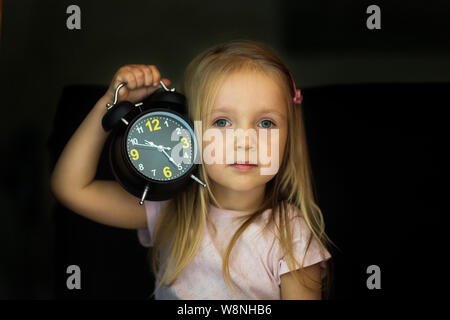 Back to school concept. A schoolgirl with an alarm clock in hand is shocked. A small girl is surprised and looking up with a black clock with a hand h Stock Photo