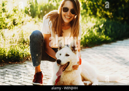 Cheerful female in casual clothes and sunglasses sitting on hunkers and hugging happy white dog lying on pavement on blurred backlit background Stock Photo