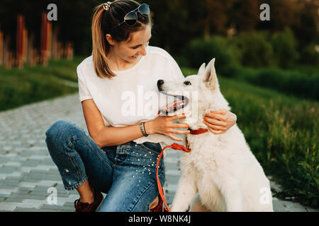 Female in casual clothes and white dog sitting together on pavement in park and hugging looking at each other Stock Photo