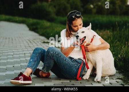 Woman in summer clothes sitting on pavement with crossed legs hugging and kissing happy white dog with open jaws looking at camera Stock Photo