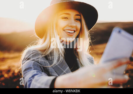 Charming young lady in trendy outfit smiling and posing for selfie on blurred background of nature on sunny day.Stylish woman taking selfie in country Stock Photo