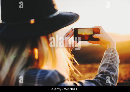 Back view of young female using smartphone to take pictures of setting sun in wonderful countryside.Anonymous woman taking pictures of sunset Stock Photo