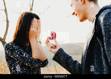 Side view of handsome young male smiling and holding small ring box while proposing to anonymous woman covering her face with hands Stock Photo