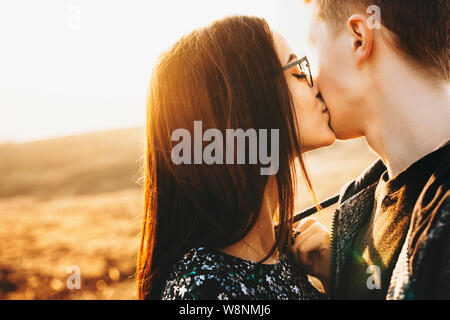 Attractive young lady in trendy glasses kissing anonymous boyfriend in cheek while standing on blurred background of countryside on sunny day.Woman ki Stock Photo