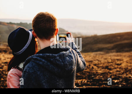 Back view of young man and woman using smartphone†to take photos of field and sunset sky in evening.Anonymous couple taking pictures on sunset Stock Photo