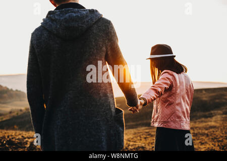 Back view of young lady holding hand of boyfriend and leading him in countryside field against sunset sky.Anonymous couple walking in meadow in evenin