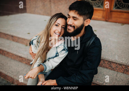 Happy bearded man and cheerful woman embracing while sitting on steps on street Stock Photo