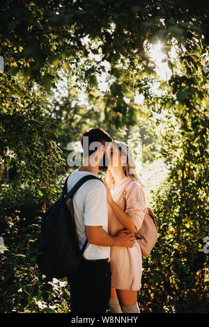 Side view of romantic young couple with backpack standing in green sunny garden and kissing happily Stock Photo