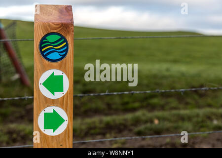 Close up of a wooden post with green arrows pointing in the direction of  the Fife Coastal Path with the logo sign above Stock Photo