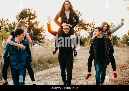 Three young men giving piggyback ride to lovely girlfriends while walking along countryside path together. Couples having fun in nature Stock Photo