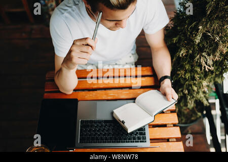 From above shot of young man in casual outfit looking at notes in notepad and thinking while sitting at table near laptop and tablet in outdoor cafe Stock Photo