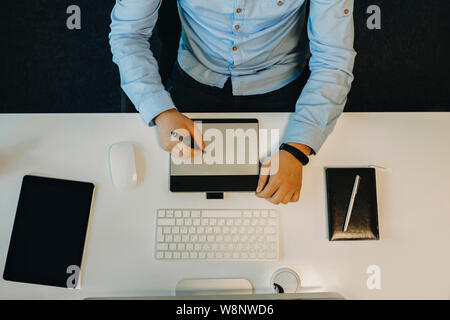 Crop view of male in elegant blue shirt sitting at white table with computer and notebook and using drawing tablet from above Stock Photo
