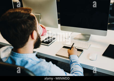 Unrecognizable man enjoying fresh hot beverage and drawing on graphic tablet near monitor with blank screen while working in office Stock Photo