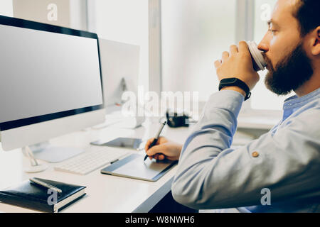 Crop bearded guy sipping hot drink from cup and using drawing tablet while working on design project in office.Bearded designer with hot drink using g Stock Photo