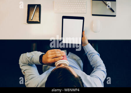 From above shot of unrecognizable male holding tablet with blank display and sipping hot drink while sitting at desk in office.Anonymous man with tabl Stock Photo