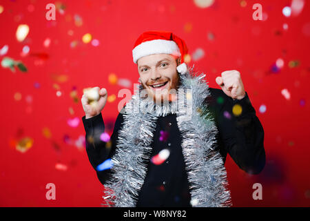 Handsome man in Christmas hat gesturing with both hands and looking at camera while celebrating success and standing behind falling confetti on red ba Stock Photo