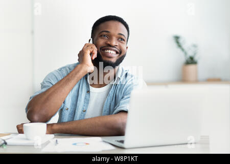 African man talking on phone, working on laptop in office Stock Photo