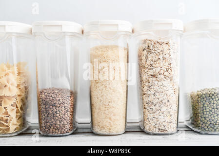 Various uncooked cereals, grains, beans and pasta for healthy cooking in plastic containers on wooden table, blue background, close-up. Food storage c Stock Photo