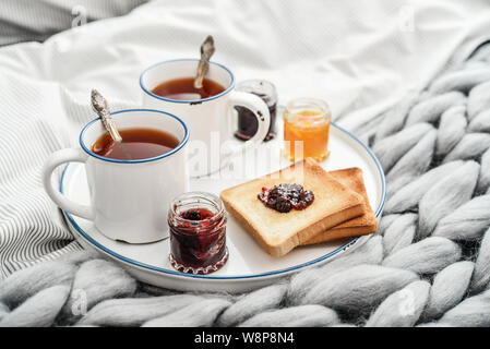 Tray with two cups of black  tea, different jam in jars and toasts on bed at breakfast time closeup Stock Photo