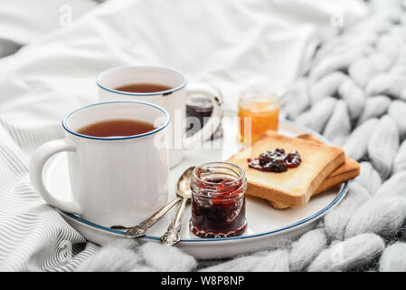 Tray with two cups of black  tea, different jam in jars and toasts on bed at breakfast time closeup Stock Photo