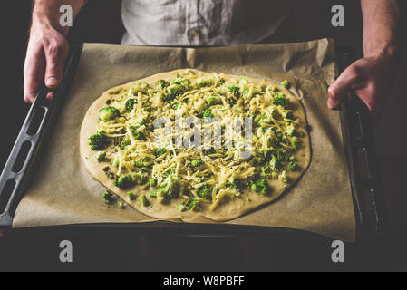 Man holding baking dish with uncooked pizza with broccoli, pesto sauce, spices and cheese Stock Photo