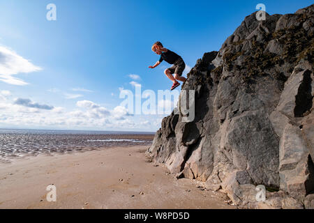 A young boy jumps from a high rock on to the beach near Sandyhills, Dumfries and Galloway showing his skill in climbing and daring. Stock Photo