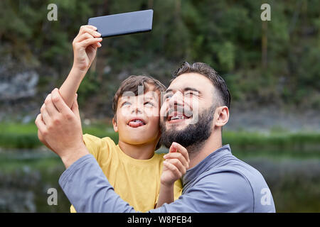 Young caucasian bearded man and his little son about 8 years old are spending fathers day on the nature near river and making faces while taking selfi Stock Photo