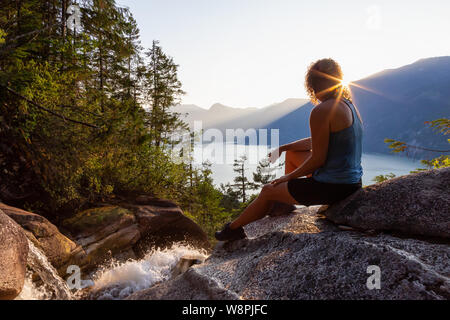 Adventurous Girl is sitting on top of a Beautiful Waterfall, Shannon Falls, and watching the sunset. Taken near Squamish, North of Vancouver, British Stock Photo