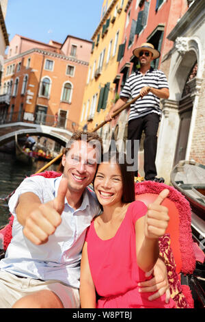 Travel. Happy couple traveling in Venice gondola giving thumbs up hand sign excited looking at camera. Romantic young beautiful couple on vacation holidays sailing in venetian canal in gondole. Italy Stock Photo