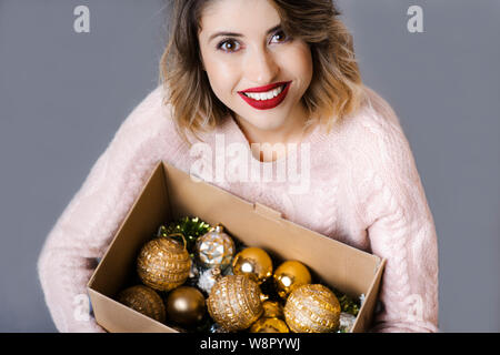 From above view of young attractive woman holding cardboard box with Christmas shiny ornaments inside and looking at camera on gray background Stock Photo