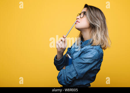 Side view of attractive young thoughtful lady in jean shirt with notepad and pen near lips on beige background Stock Photo