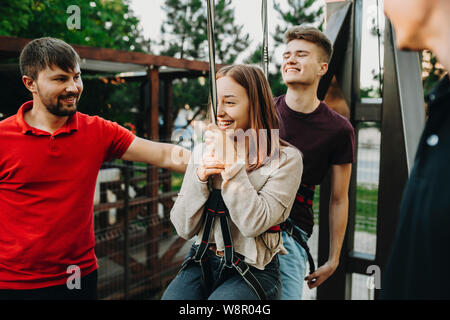 Happy young man woman in safety harness equipment hanging and preparing for ride of zip line with staff men around Stock Photo