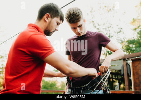 Adult staff man helping young guy with wearing safety harness gear standing on terrace in summer park Stock Photo