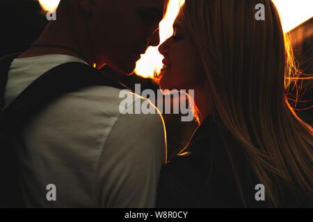 Crop back view of beautiful young couple touching noses with closed eyes standing outdoors on blurred backlit background of setting sun Stock Photo