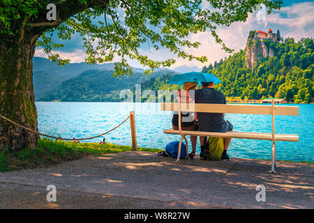 Adult romantic couple sitting and embracing on the bench in the rain with umbrella. Carefree couple in love enjoying the lake and castle. Happy love c Stock Photo