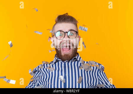 Joyful young hipster male in a glasses is laughing happily among flying confetti on a yellow background. The concept of a mega party and holiday. Stock Photo