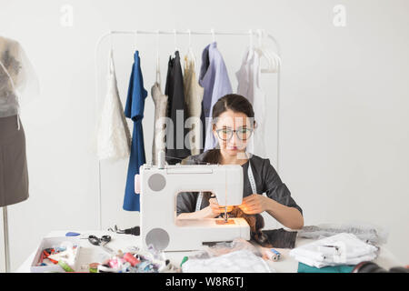 Dressmaker, tailor and fashion concept - Smiling female fashion designer using sewing machine and sitting behind her desk Stock Photo