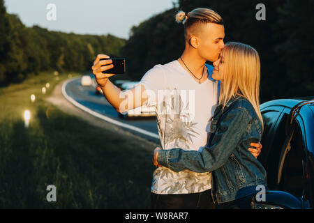 Handsome young man kissing lovely woman in forehead and using smartphone to take selfie while standing on roadside in countryside Stock Photo