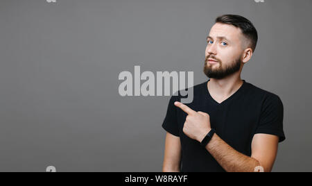 Attractive bearded guy in black T-shirt looking at camera and pointing aside while standing on gray background Stock Photo
