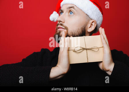 Handsome male in Christmas hat looking away and thinking while standing on bright red background and holding lovely present Stock Photo