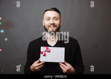 Handsome young man with lovely gift box smiling and looking at camera while standing on gray background under falling confetti Stock Photo
